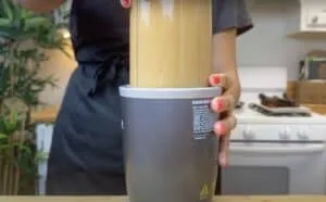 Person blending a smoothie in a kitchen with a countertop blender, wearing a dark apron.